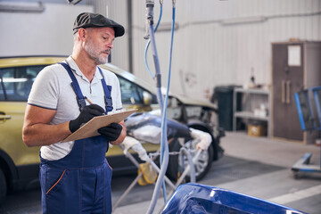 Bearded auto mechanic writing on clipboard at vehicle repair shop