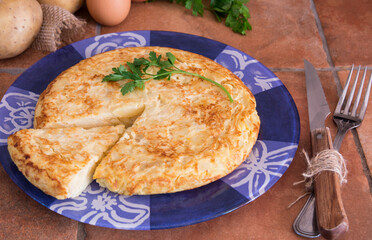 Spanish omelette made with potatoes, eggs and onion, with a piece of parsley on top, on a blue plate in a rustic kitchen 