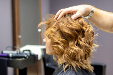 Side view of male hairdresser checks curly hairstyle of young caucasian woman in hair salon. Soft...