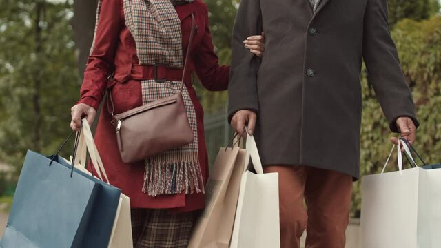 Travelling up shot of cheerful elderly Caucasian family couple dressed elegantly walking down street after shopping, talking, laughing, holding multiple paper bags