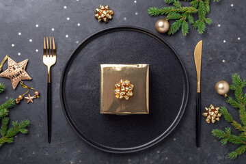 Christmas table setting with black ceramic plate, gift box and gold accessories on black stone background. Top view. Copy space