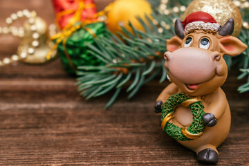 Toy bull in a Santa Claus hat on a dark wooden background.Christmas tree branch with little gift boxes and balls in the background.
Bull as a symbol of the New year 2021.New Year,Christmas concept.