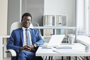 Fototapeta premium Portrait of African businessman in suit sitting on chair at his workplace and looking at camera at office