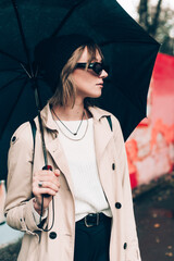 young stylish woman wearing long beige coat, white boots, black hat, umbrella and backpack posing...