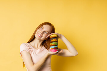 Happy joyful redhead woman sweet tooth holding pile of colorful glazed donuts, broadly smiling looking at camera standing against yellow studio wall. Pastry, bakery concept.