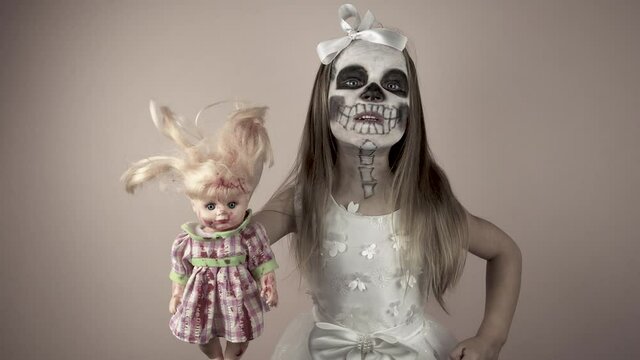 A little girl in a white dress with a painted face holds a bloody doll in her hands. She shows her teeth and growls. A terrible image of a skull on the face. 4k