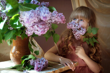 a little blonde girl sits at a table with a bouquet of lilacs in a beautiful dress and sniffs the flowers