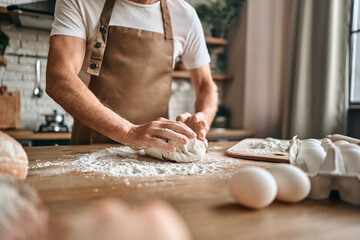 Man in apron holding the dough on a wooden table adding flour to it