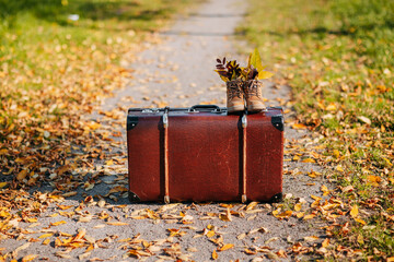 Old brown boots with leaves inside. Boots and brown vintage suitcase in autumn forest. Path with yellow leaves. Leather suitcase under the tree. Autumn nature, yellow and red foliage.