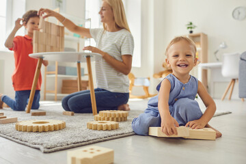 Cute little kid looking at camera and smiling while elder brother and mum are playing in background