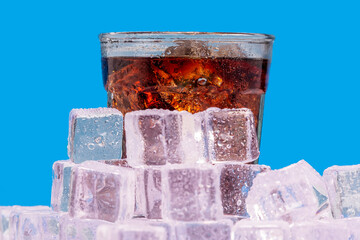Ice cold glass of soda, pop, soda pop, cola, fizzy drink, carbonated beverage sits surrounded by ice cold ice cubes with condensation against a blue wall looking refreshing and inviting.