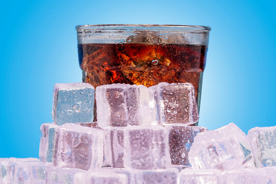 Ice cold glass of soda, pop, soda pop, cola, fizzy drink, carbonated beverage sits surrounded by ice cold ice cubes with condensation against a blue wall looking refreshing and inviting.