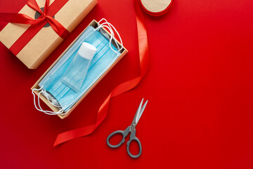 Packing a Christmas present during Coronavirus epidemic. Face masks and hydroalcoholic gel inside a cardboard box decorated with a bow on a red background.
