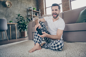Full body photo of content guy sit floor carpet enjoy playing video games joystick wear checkered plaid pants trousers white t-shirt in house indoors