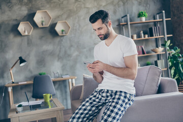 Photo of concentrated man sit divan use smartphone search coronavirus information wear checkered plaid pants trousers white t-shirt in living room house indoors