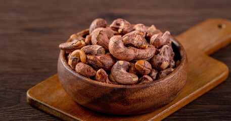 Cashew nuts with peel in a wooden bowl on wooden tray and table background, healthy raw food plate.