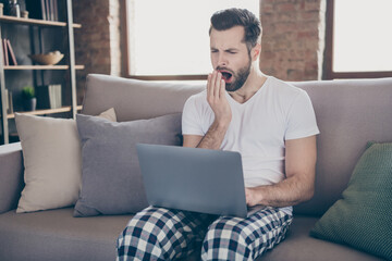 Photo of exhausted man sit divan work remote laptop tired prepare startup progress innovation report yawn mouth hand wear sleepwear nightwear in house room indoors