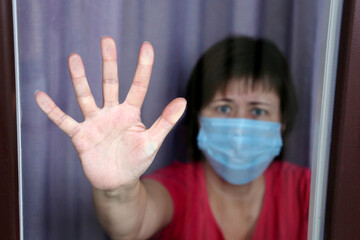 Quarantine during the covid-19 coronavirus epidemic, female palm of hand on the window. Worried woman in a medical face mask looks through the glass, plea of help