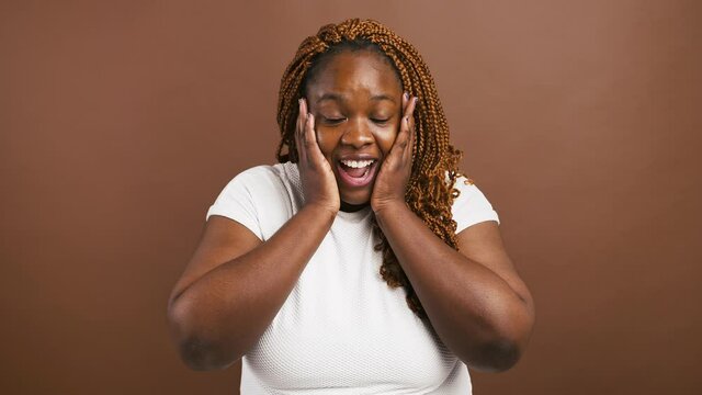 Surprised african american female touching her cheeks in amazement, brown background, slow motion