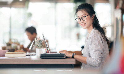 Fototapeta premium young beautiful asian woman woking on tablet computer, smiling and looking at camera while sitting at the table in co-workspace.