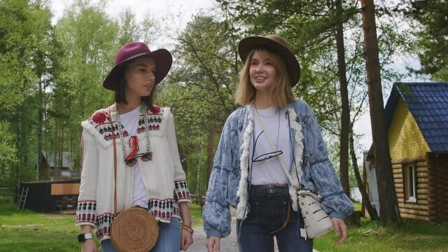 Front-view medium footage of attractive young women dressed up in boho chic style clothes walking among trees talking and smiling to music festival