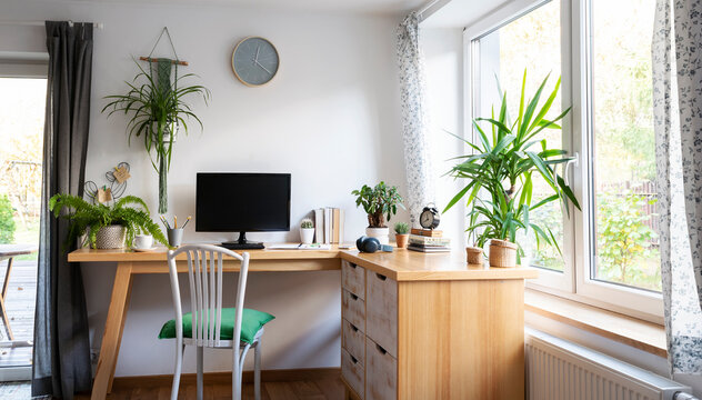 Interior of office room with big window, green plants, wooden furniture and computer with blank monitor with mock up. Light workspace with desk in vintage style. Home office. Banner.