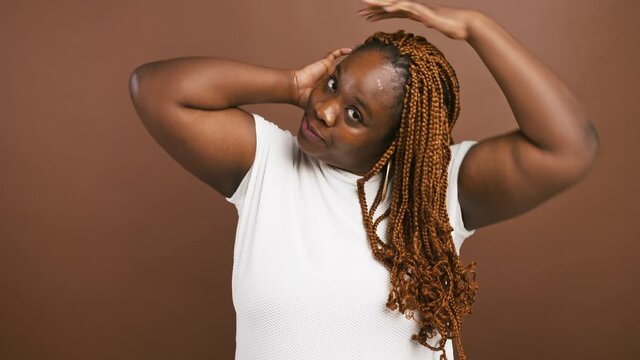 Young african american woman fixing her hair, enjoying her appearance, smiling at camera, brown studio background