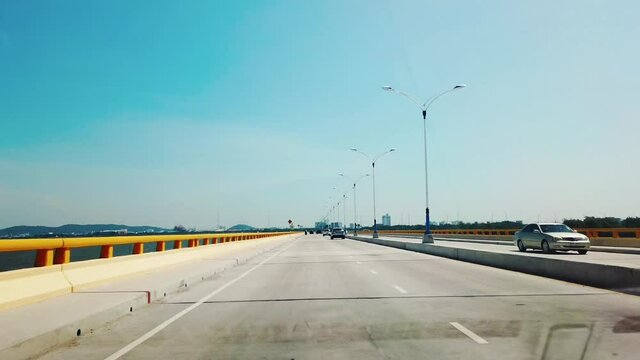 The footage from the front camera of a car being driven slow down over the speed bump on the road along the sea in the bright blue sky on a sunny day at Chonburi Province on the country of Thailand
