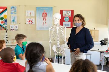 Female teacher wearing face mask using human skeleton model to teach students in class