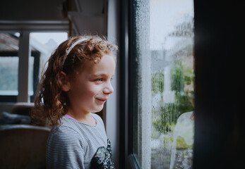 Happy girl with cute smile enjoying looking at rain from window at home
