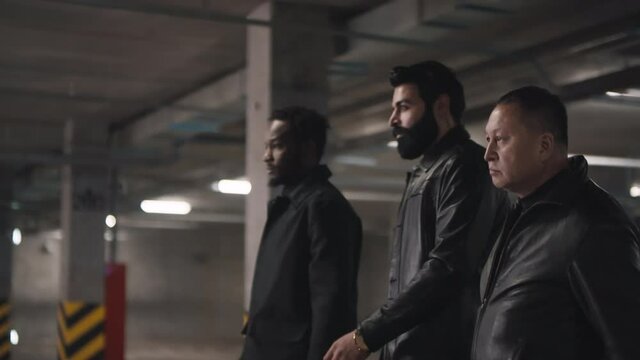 Side-view through columns footage of Eastern and Western assertive gang members walking down underground parking going on deal