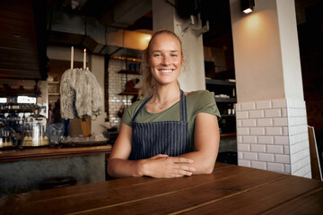 Portrait of smiling young female waitress wearing apron standing leaning on table in cafe