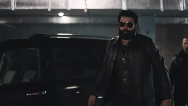 Tilting up footage of brutal bearded man in sunglasses getting out of big black car at underground parking with squad of criminals going on deal