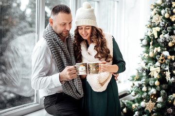 Christmas. Love. Home. Young couple in warm clothes is holding cups, talking and laughing while sitting at home near the Christmas tree