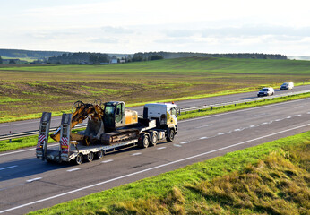 Trailer truck with long platform transport the Excavator on highway. Earth mover backhoe on heavy...
