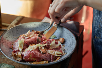 A female hand is preparing a vegetable salad with beef roast beef. A plate of salad with beef roast beef, onions, arugula, pepper and tomatoes on a black table. Top view.