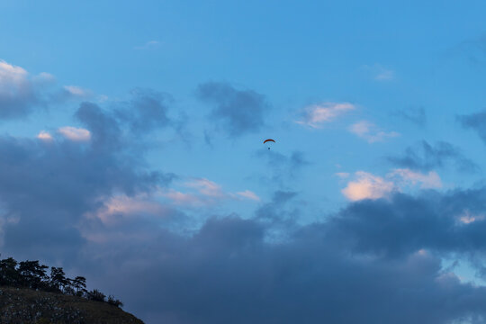 Blue sky with clouds. There is a small rogalo in the sky in the distance. On the left is a wooded hilltop.