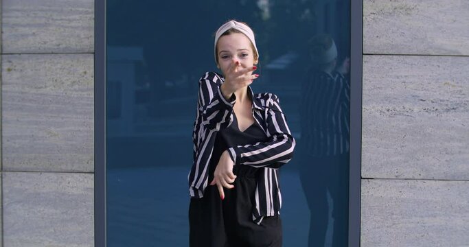 Young active cheerful student girl wears headband and striped jacket actively moves her arms posing in front of camera stands outdoors woman dances on street shaking her body, concept of enjoying life
