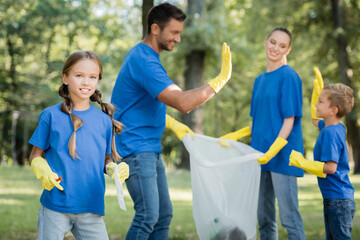 girl looking at camera while parents holding recycled bag and giving high five on blurred background, ecology concept