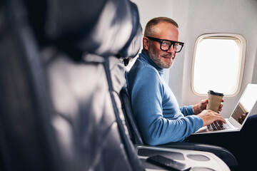Naklejka na ściany i meble Pleased businessman in eyeglasses sitting in the aircraft cabin