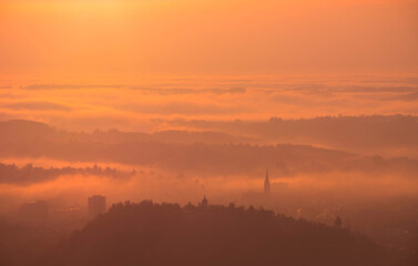Amazing foggy sunrise over the city of Graz with Schlossberg hill and Church of the Sacred Heart of Jesus tower, in Styria region, Austria. Panoramic view from Plabutsch mountain on autumn morning.