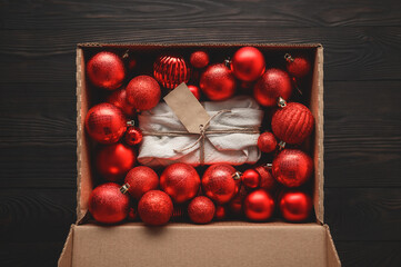 A Christmas present, wrapped in natural textile material, lies among the Xmas balls in a cardboard box. Dark brown wooden table. Zero waste concept. View from above.