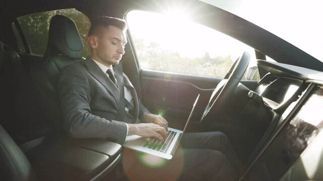 Self-driving electric car autopilot demanding driver attention to hold steering wheel and take control on highway. Businessman working on laptop at sunset.