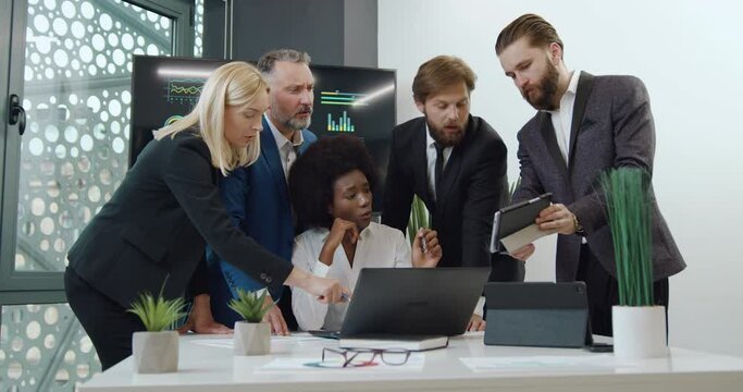 Beautiful experienced adult black-skinned female boss sitting at the table and listening the versions to improve results of joint work from her confident subordinates standing ner her in office