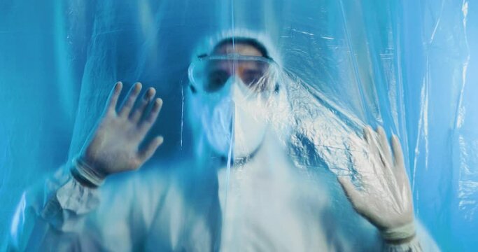 Medico o infermiere con tuta bianco covid si mette le mani in testa dalla disperazione dietro a un telo di plastica trasparente in ospedale.