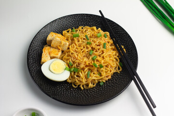 Korean Spicy instant fried noodle served on black textured ceramic plate, topping with half of boiled egg, tofu, and spring onions. Isolated white background. 
