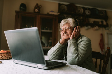 Cute caucasian female senior with glasses and headphones on her head while sitting at a table in front of a laptop and talking on a video call with her family during quarantine COVID - 19 coronavirus
