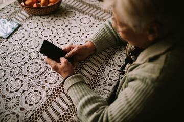 Fototapeta na wymiar Close up of a caucasian senior woman's hand while sitting at a kitchen table and holding a phone in her hand, she calls her family during quarantine COVID - 19 coronavirus