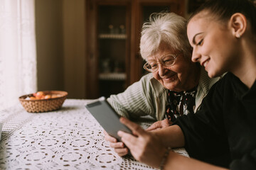 A cute gray-haired retired grandmother and her teenage caucasian granddaughter look at a tablet as they sit at a table in their grandmother's house. Grandma's education about new technology - obrazy, fototapety, plakaty