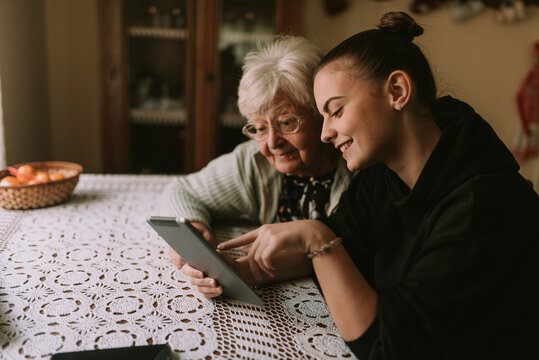A smiling caucasian grandmother and her teenage granddaughter look at a tablet while sitting at a table in the kitchen. The granddaughter teaches her grandmother to use new technology